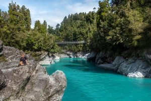 Nathan from Backyard Travel Family sits on a rock with the Hokitika Gorge Walk swingbridge in behind. The azure blue/green waters are glistening in the sunlight