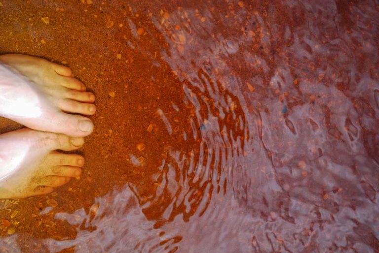 Close up of bare toes, dipped into the red, orange water at Coca Cola Lake in Doubtless Bay