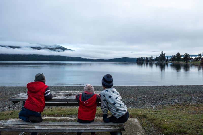 the Backyard Travel Family kids sit at a picnic table overlooking Lake Te Anau on a calm cloudy morning