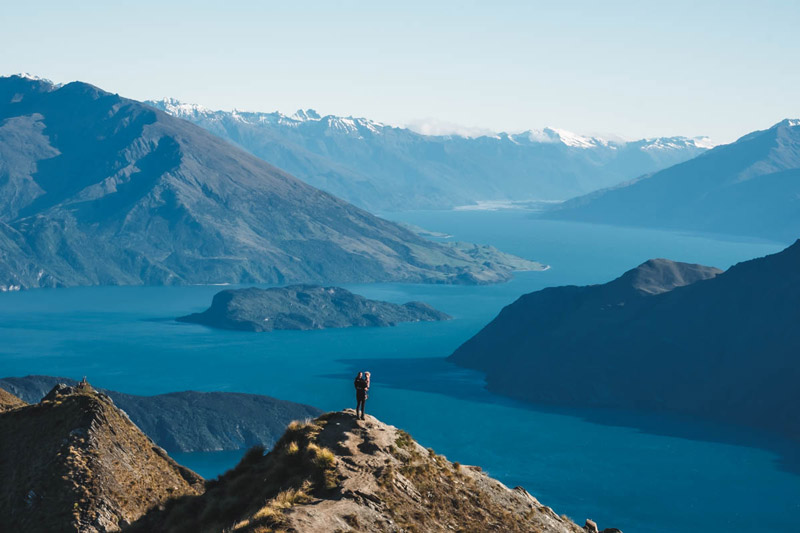 Jen and 2.5 year old Emilia stand on top of the Roys Peak viewpoint with blue lake views and mountain ranges in behind