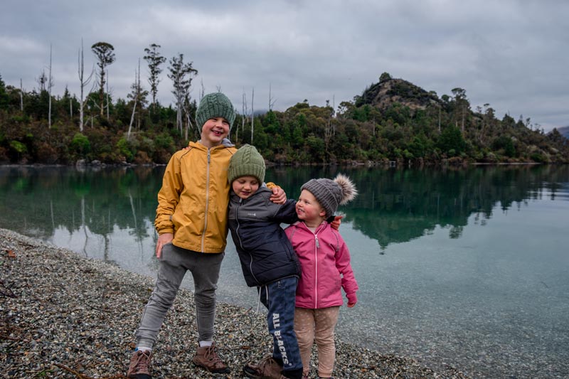 Three kids from Backyard Travel Family embrace on the beach at Bobs Cove Queenstown