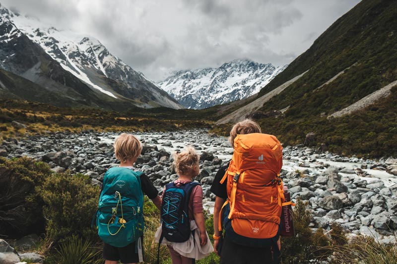 3 children with hiking packs on their back look at Mt Cook, the top covered in cloud, on the way to the Hooker Hut