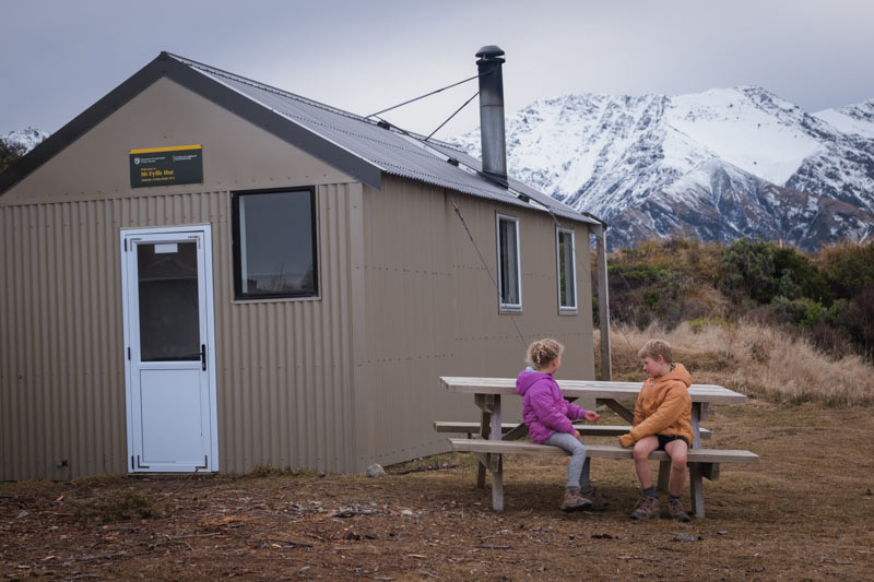 two kids sit in front of Mt fyffe Hut with snowy mountains in the background