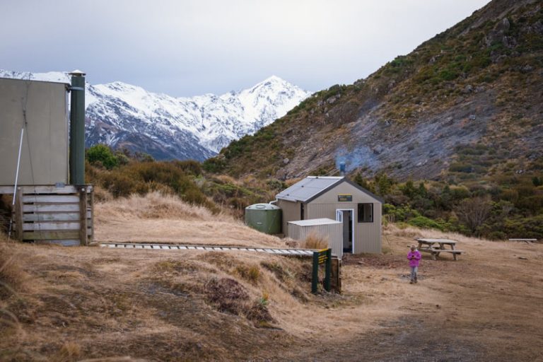 snowy mountains sit in the distance beside Mt Fyffe Hut. a young girl walks in front