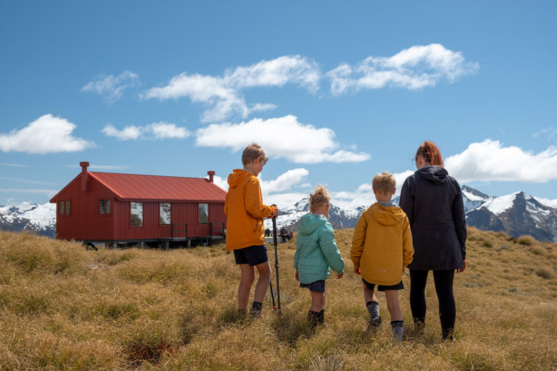 Family walk towards red Brewster Hut on a blue spring day after hiking the Brewster Track